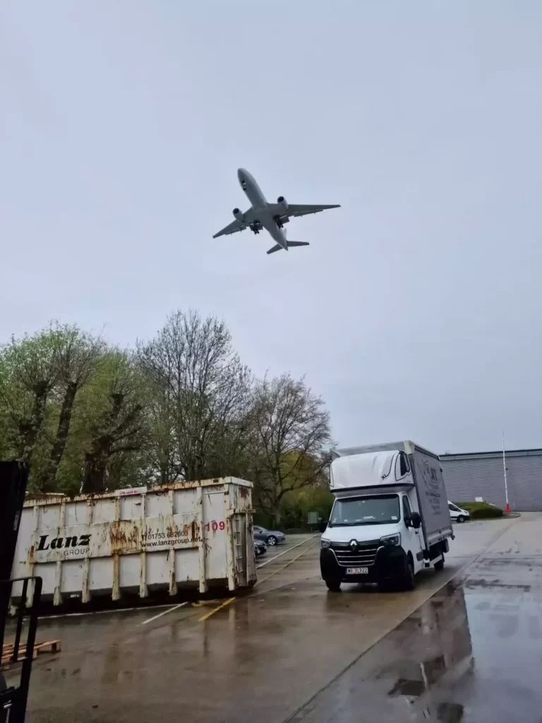 Na zdjęciu widać samochód dostawczy firmy transportowej zaparkowany na placu w deszczowy dzień, przygotowany do realizacji przeprowadzki. Nad pojazdem przelatuje samolot, co symbolicznie podkreśla możliwość organizacji przeprowadzek na dłuższych trasach, również międzynarodowych. Całość oddaje charakter usług – sprawny, terminowy i elastyczny transport mienia niezależnie od odległości.